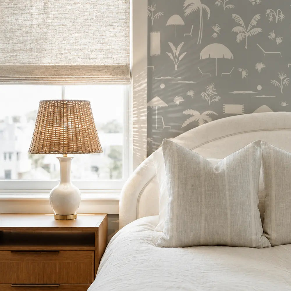 A styled bedroom corner featuring a bed with cream linen bedding and pillows, a wooden nightstand with a woven lamp, a window with a textured beige roller shade, and a gray wall with a white beach-themed patterned wallpaper.