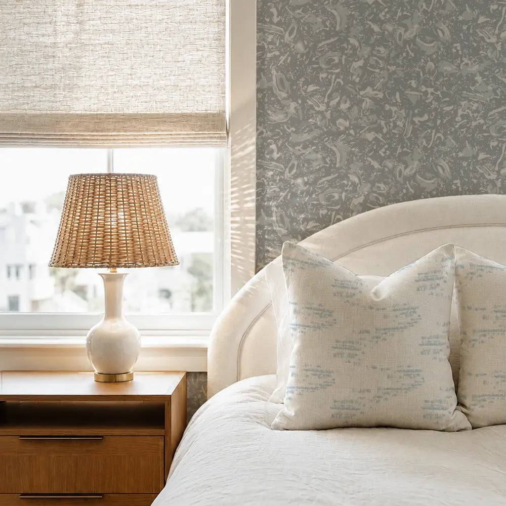 A styled bedroom corner featuring a bed with cream bedding and patterned pillows, a wooden nightstand with a lit table lamp, and a window with a textured beige roller shade. The wall behind the bed has gray floral wallpaper. Emily Daws Salt Marsh Pillow in Shallow