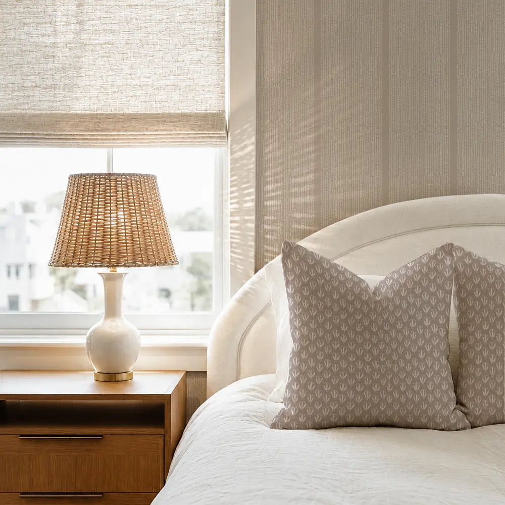 A styled bedroom corner featuring a bed with cream bedding and patterned beige pillows, a wooden nightstand with a white lamp, and a window with a woven roller shade. Kiawah Pillow in Rosewood by Emily Daws