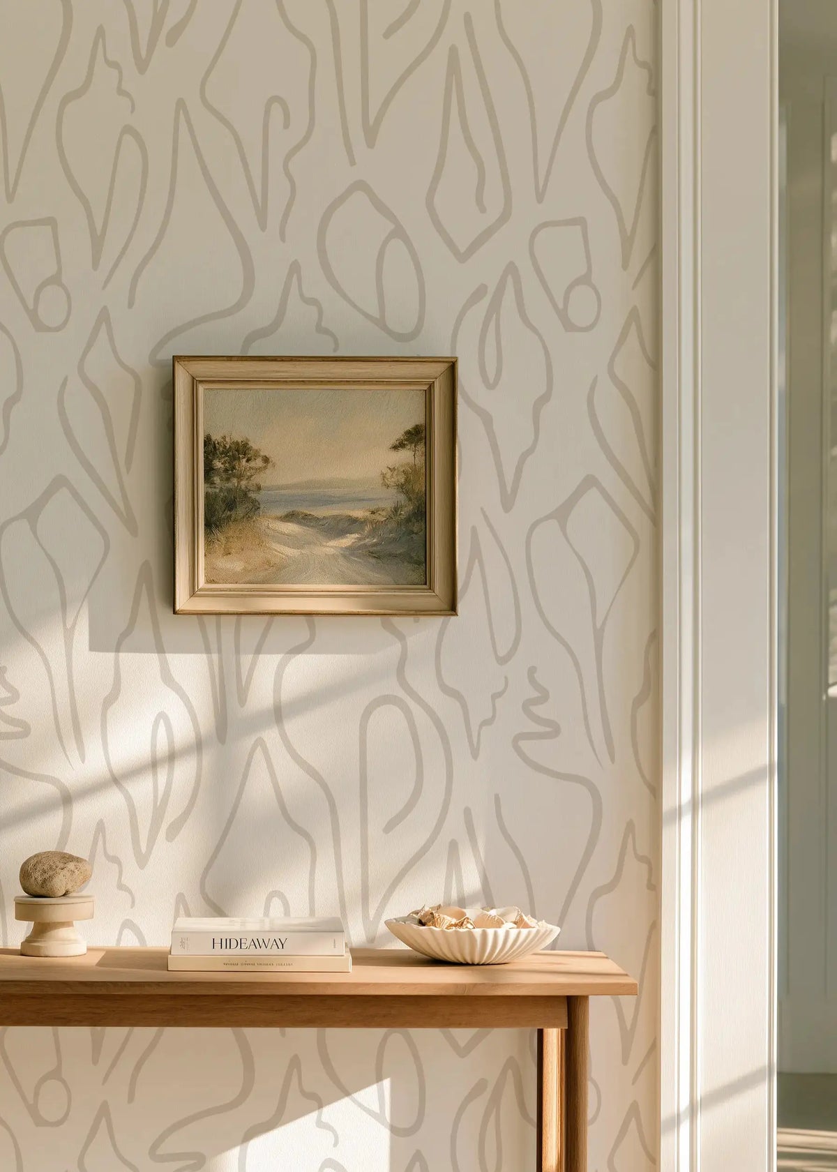 A styled interior corner featuring a small framed landscape painting hanging on a wall with abstract beige shell wallpaper by Emily Daws in Charleston. Below the painting is a light wood console table holding a decorative bowl, stacked books, and a small stone object. Edisto wallpaper in Shore.