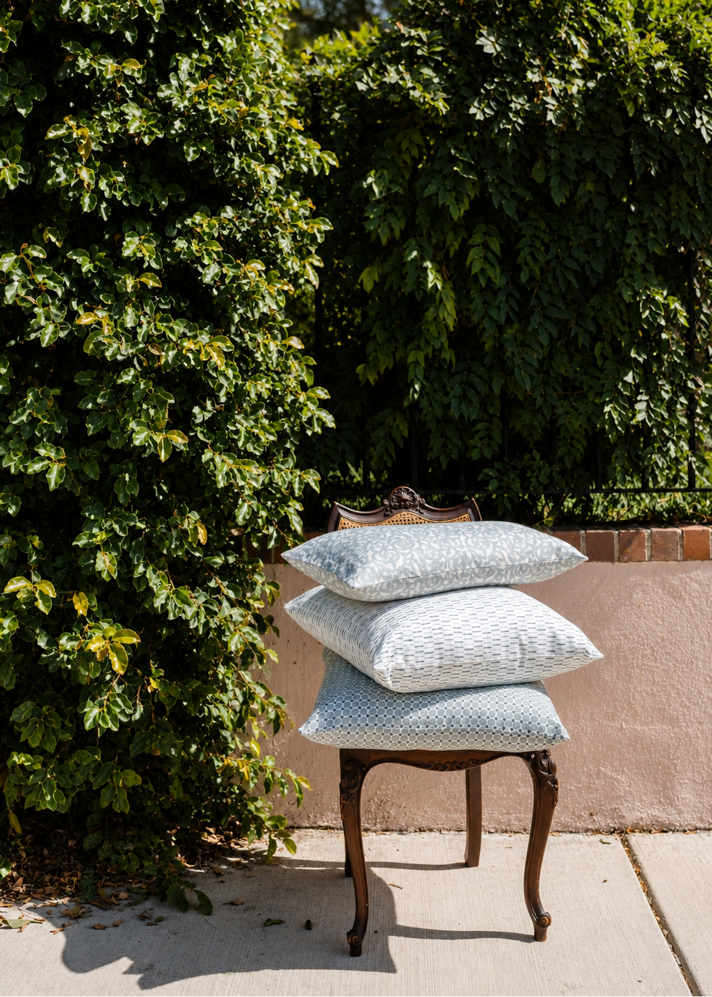 Three light blue patterned throw pillows stacked on a dark wooden chair outdoors in front of a green hedge and beige wall.
