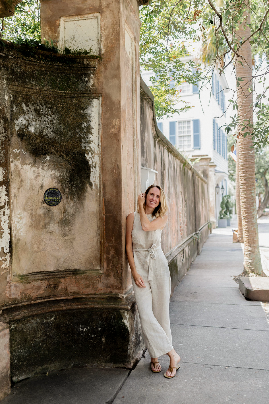 A woman wearing a light beige sleeveless jumpsuit and sandals stands on a sidewalk next to a weathered stone wall in an outdoor urban setting. Emily Daws in Charleston SC.