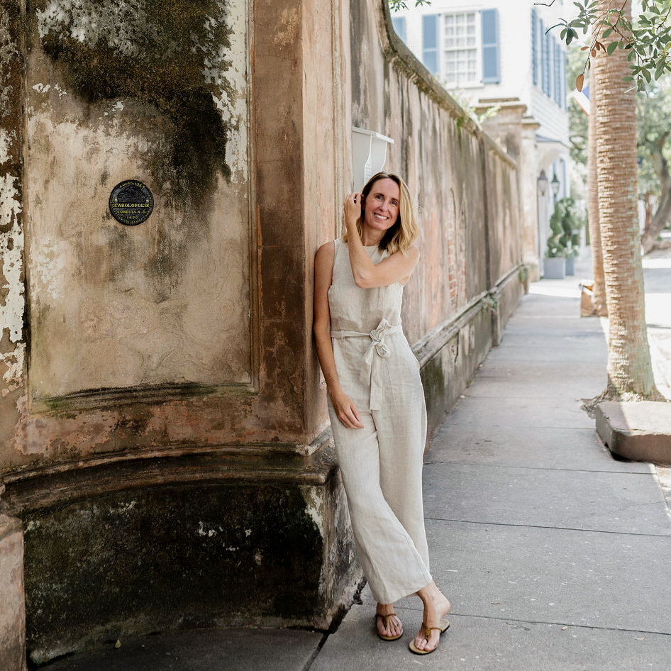A woman wearing a light beige sleeveless jumpsuit and sandals stands on a sidewalk next to a weathered stone wall in an outdoor urban setting. Emily Daws in Charleston SC.