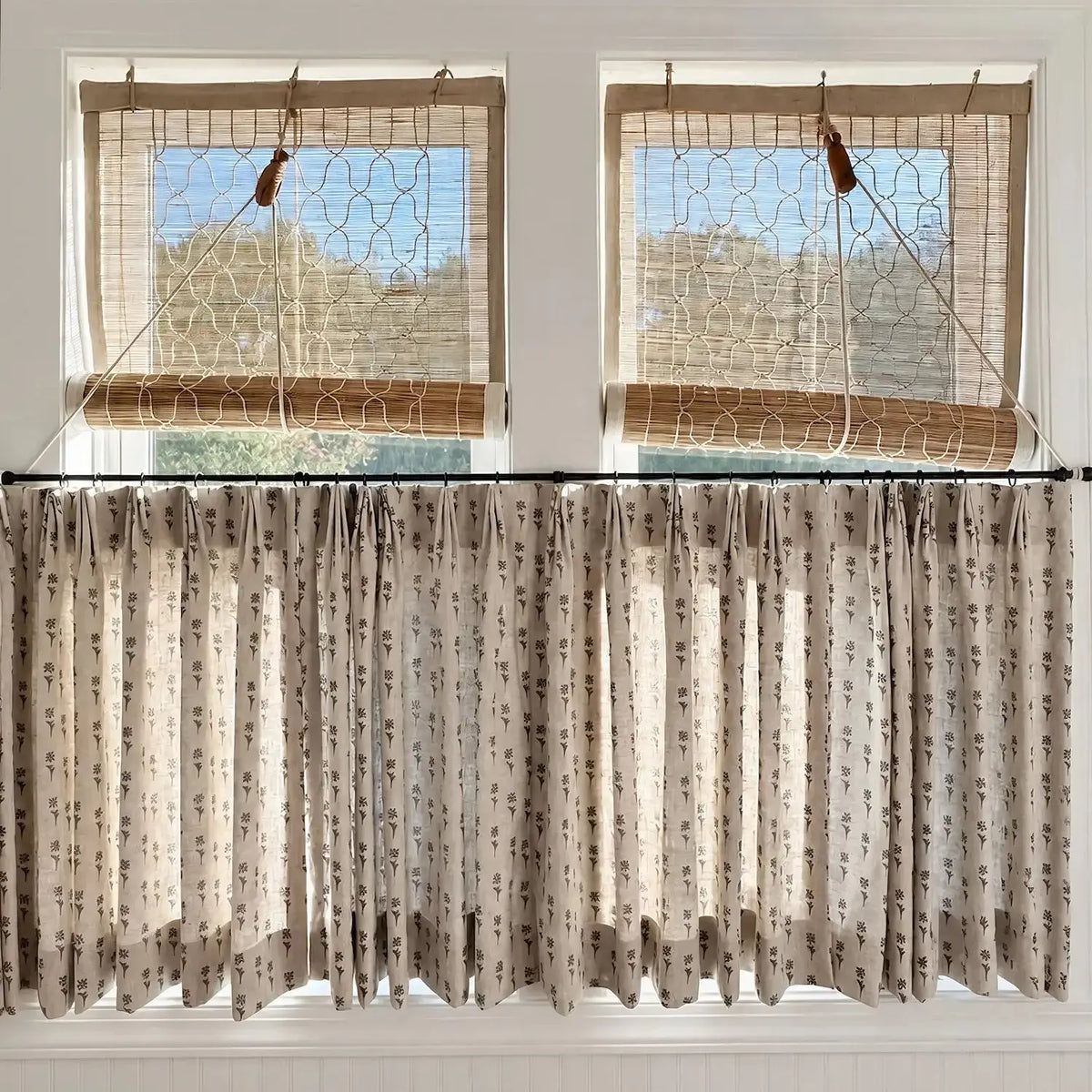 A set of beige patterned curtain panels hanging from a black rod over a window, with matching woven window blinds above on a white paneled wall.
