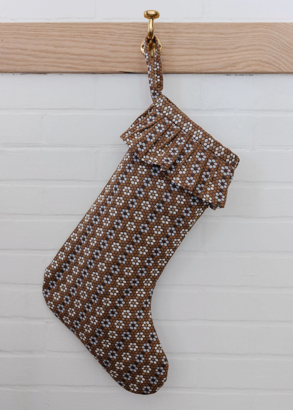 A brown woven Nectar-pattern holiday stocking hanging from a brass hook on a white shiplap wall.