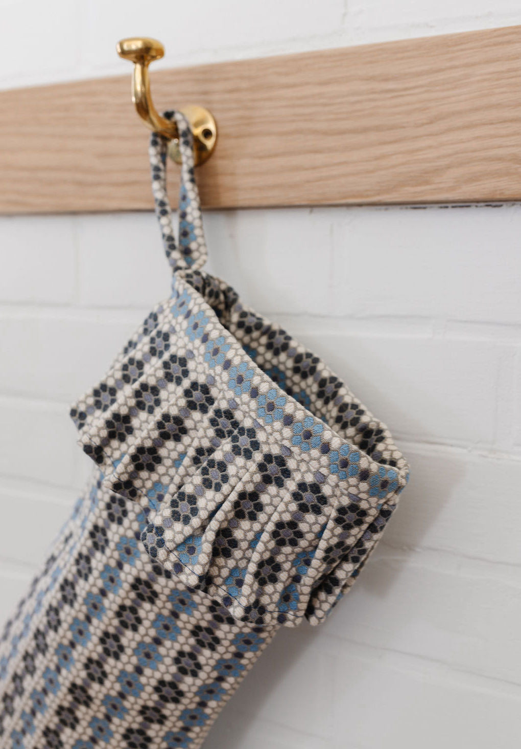 A Nectar woven holiday stocking by Emily Daws displayed on a brass hook against white shiplap.