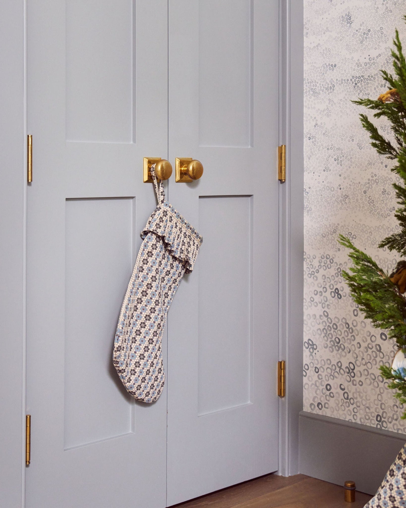 A holiday stocking by Emily Daws, hanging on double brass hooks on a white paneled door near a Christmas tree in the Southern Living Idea House.
