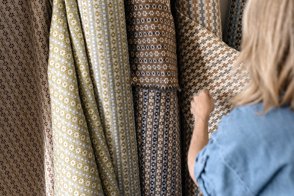 A person in a blue shirt is shown from behind, holding and examining patterned fabric rolls in a sunlit room. The fabrics feature intricate geometric and floral motifs in muted tones of beige, brown, olive green, and blue. Emily Daws Nectar upholstery fabric by the yard.