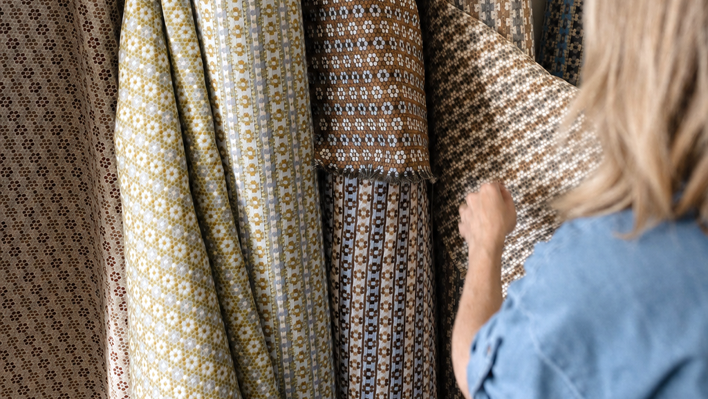 A person in a blue shirt is shown from behind, holding and examining patterned fabric rolls in a sunlit room. The fabrics feature intricate geometric and floral motifs in muted tones of beige, brown, olive green, and blue. Emily Daws Nectar upholstery fabric by the yard.