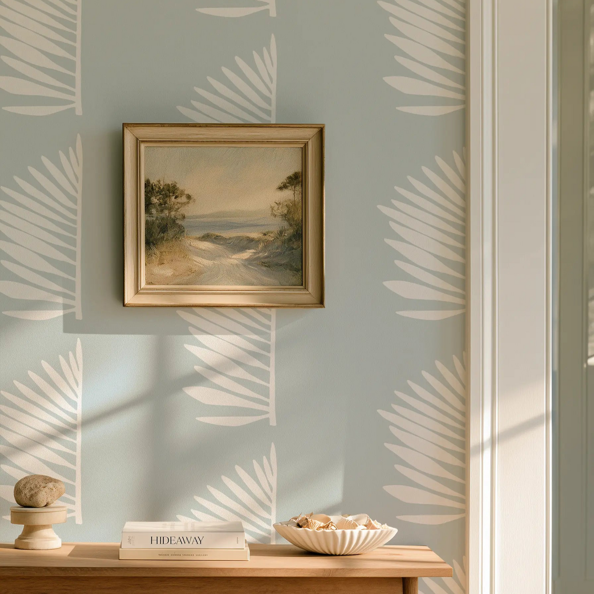 A styled interior scene with pale blue wallpaper featuring a repeating white palm leaf pattern, Palmetto by Emily Daws, a small framed landscape painting on the wall, a wooden console table, books, and a bowl of fruit in warm natural light. Palmetto wallpaper in Haint Blue, designed in Charleston.