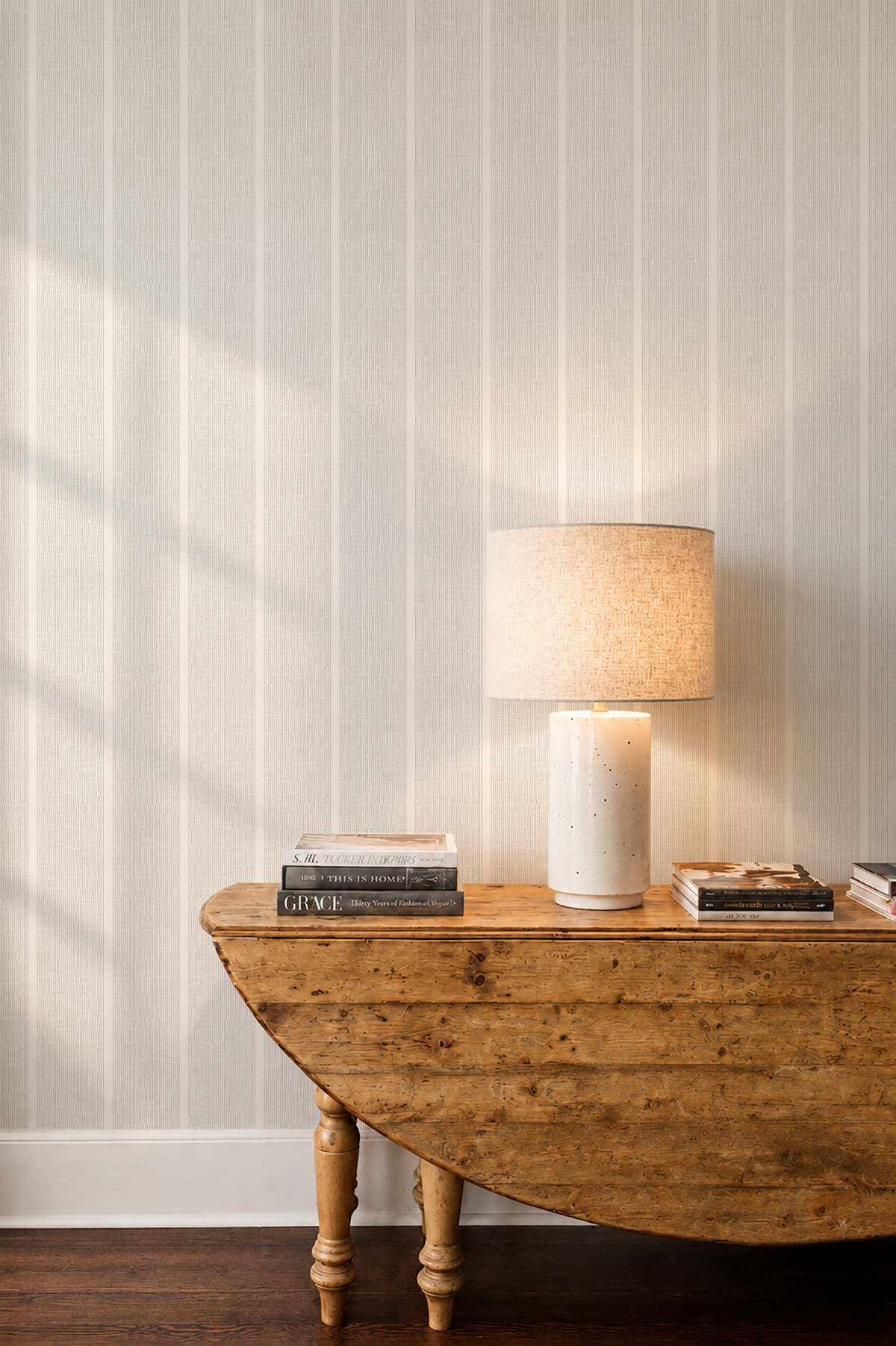 A warm, modern interior scene featuring a white ceramic table lamp with a textured beige shade resting on a rustic wooden table against a white wall with vertical paneling. Emily Daws Sandbar Wallpaper in Driftwood