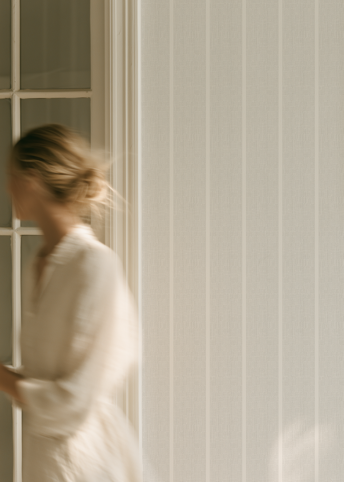 A woman in a flowing white dress walks past a window with white paneled trim, set against a wall with vertical white stripes and warm natural light. Emily Daws Sandbar wallpaper in mist.