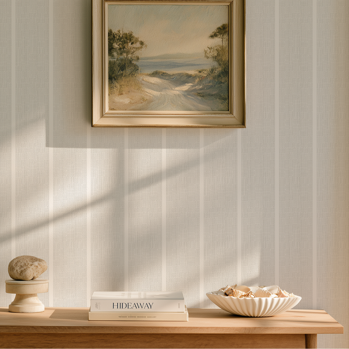 A framed landscape painting hangs on a vertically paneled wall above a wooden console table styled with books, a bowl of shells, and a small decorative stone in a softly lit interior. Emily Daws Sandbar wallpaper in Mist