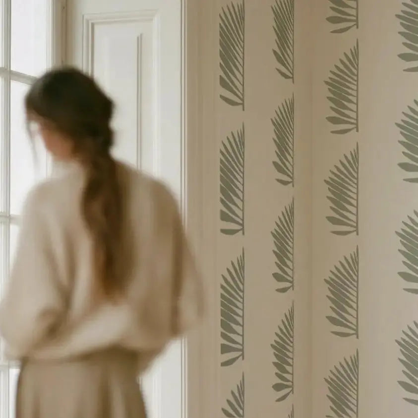 A woman stands in a softly lit room with a blurred silhouette on the left, facing a wall covered in repeating vertical palm-leaf wallpaper by Emily Daws in muted sage green on a warm off-white background.