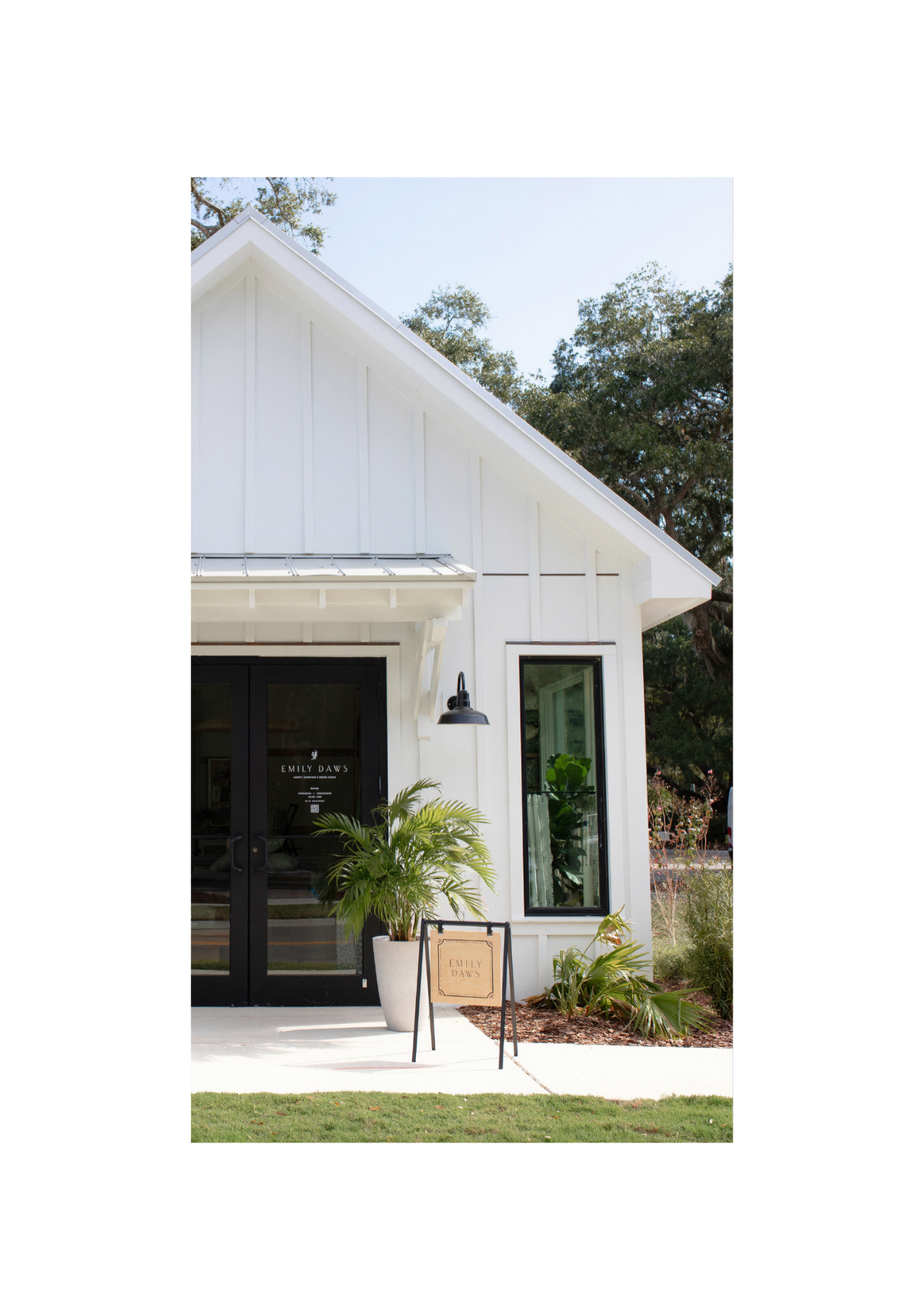 A white single-story house exterior with a dark front door, large windows, a small black hanging light fixture, a black-and-white A-frame sign, and green plants and trees around the property. Emily Daws Showroom