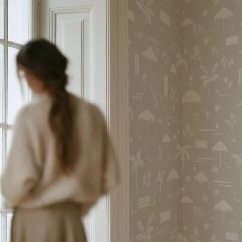 A woman in a cream-colored outfit stands in profile near a window, looking out while the wallpaper by Emily Daws on the wall features a white tropical beach-themed pattern with palm trees, umbrellas, and lounge chairs. Folly Toile wallpaper in Sand.