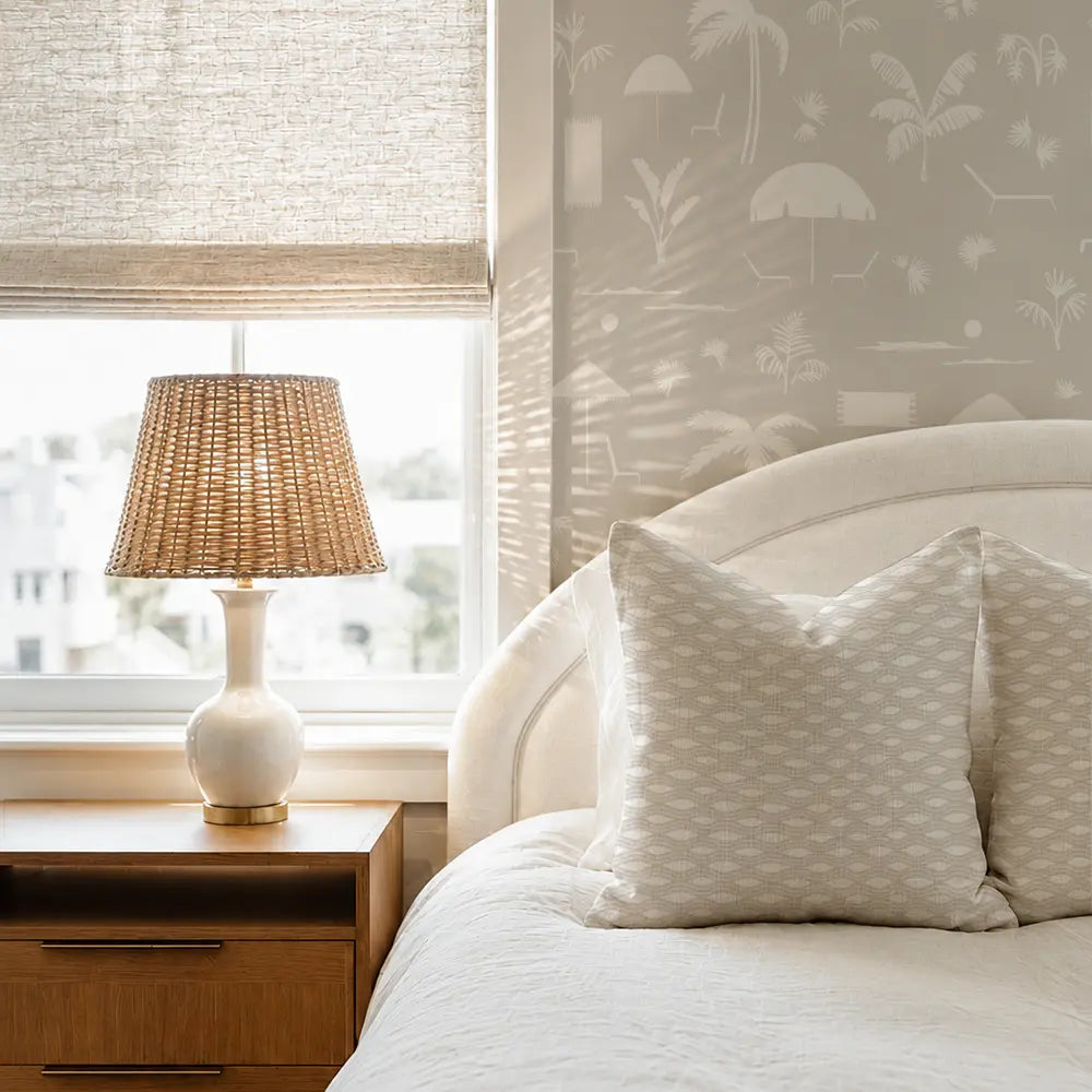 A styled bedroom corner featuring a bed with white textured bedding and pillows, a wooden nightstand with a woven lamp, and a window with a beige roller shade. The wall behind the bed has a light gray wallpaper with a white beach umbrella and palm tree pattern. Emily Daws Inlet Pillow in Beach