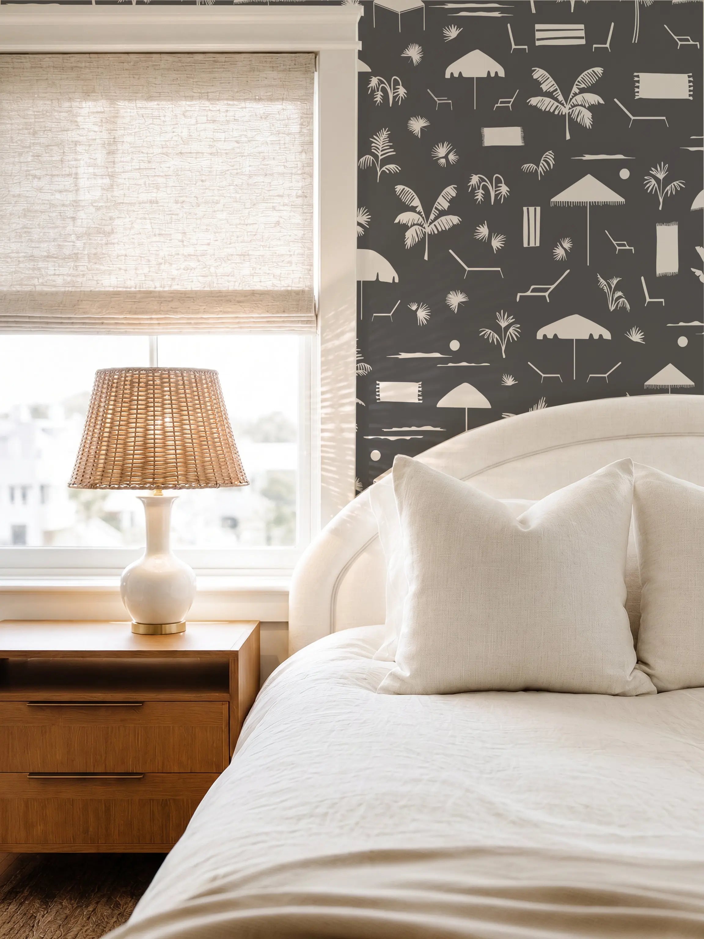 A styled bedroom corner featuring a bed with white linens and pillows, a wooden nightstand with a woven lamp, and a dark blue coastal wallpaper by Emily Daws with a white beach-themed pattern of umbrellas, palm trees, and lounge chairs.