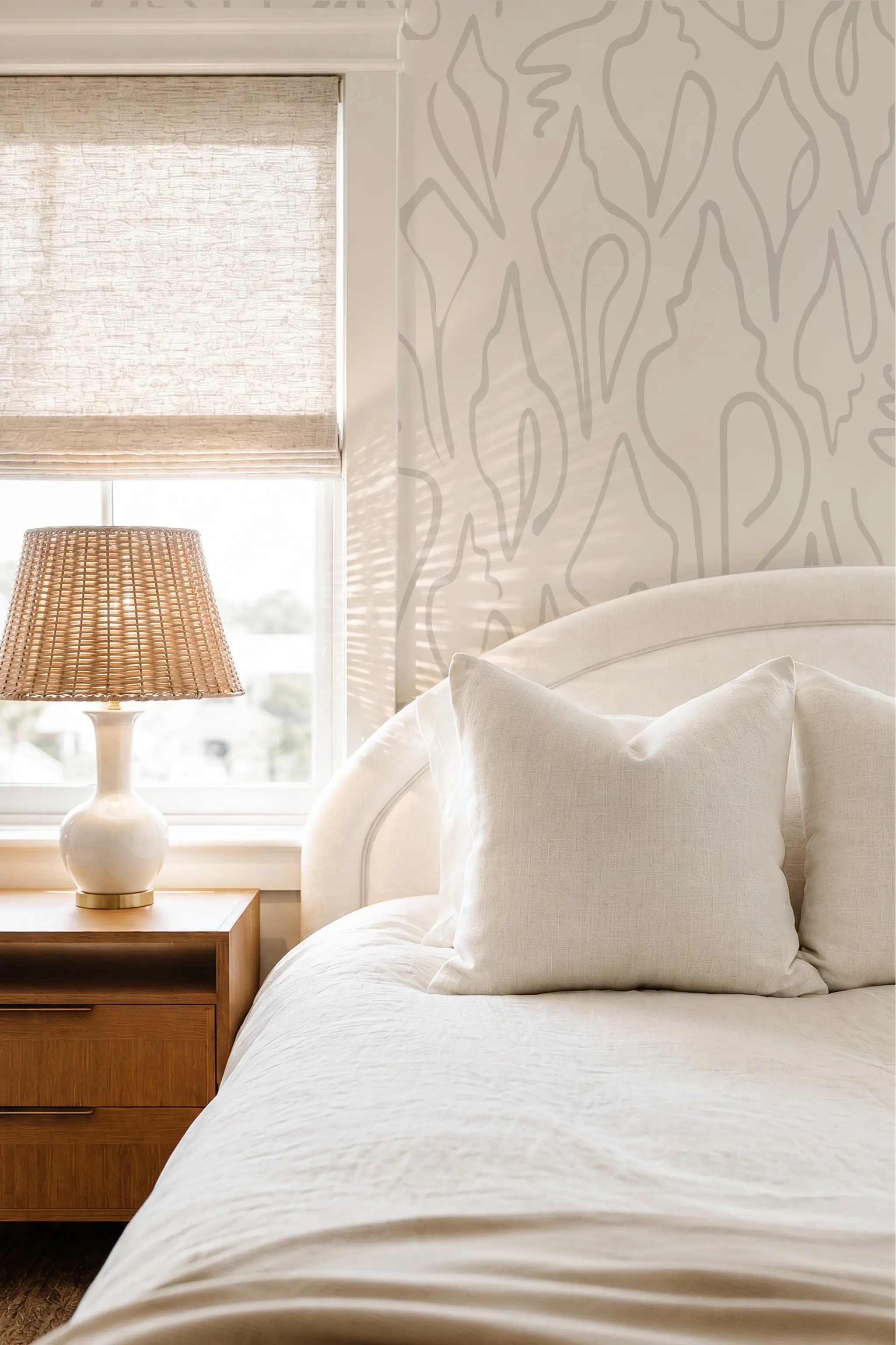 A styled bedroom corner featuring a white upholstered bed with cream bedding and pillows, a wooden nightstand with a woven lamp, and a window with a beige roller shade. The wall behind the bed has a light coastal neutral shell pattern wallpaper by Emily Daws.
