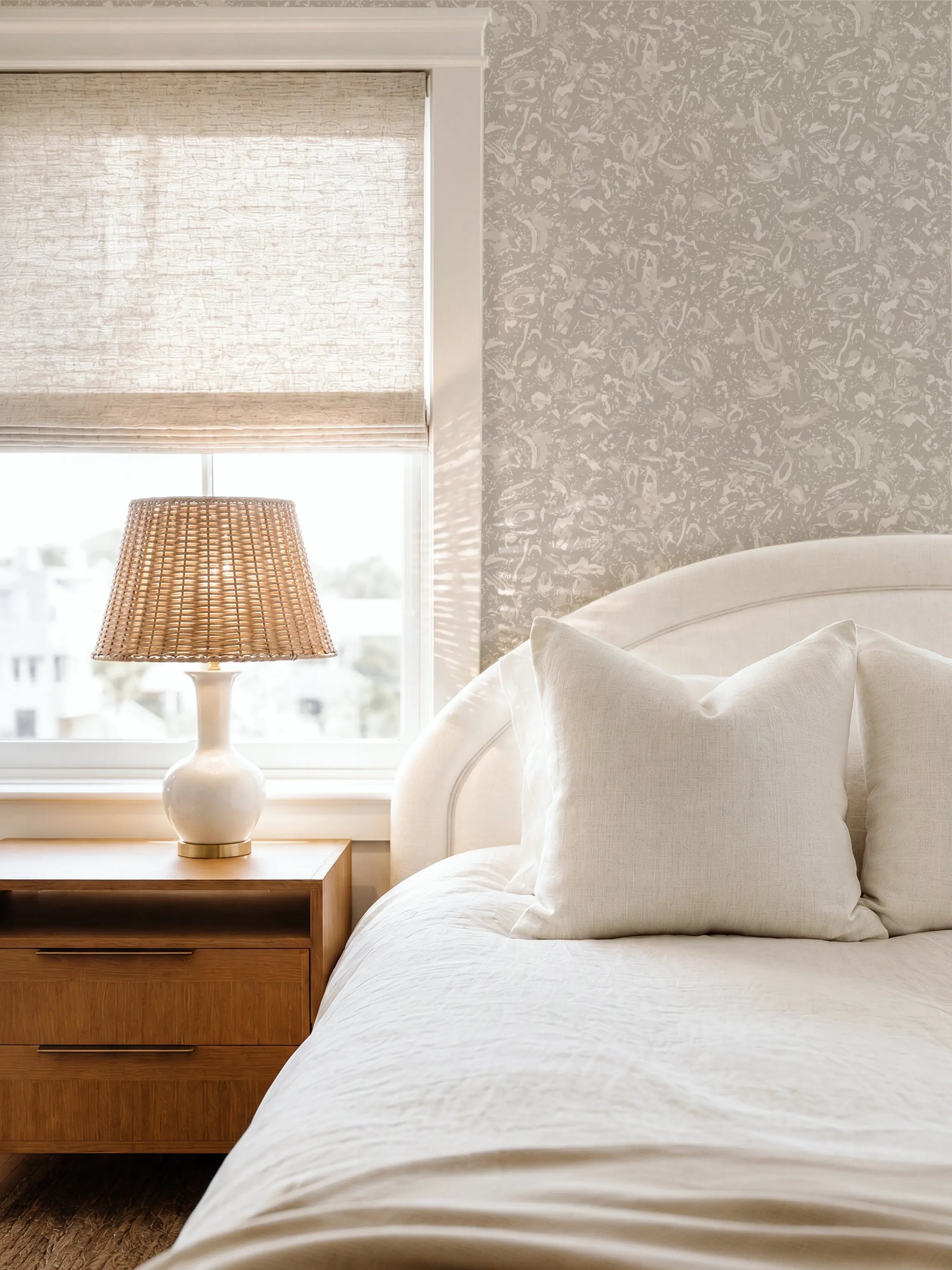 A styled bedroom corner featuring a white upholstered bed with cream bedding, a wooden nightstand, a woven rattan table lamp. The wall behind the bed has textured coastal wallpaper by Emily Daws.  River Reef wallpaper in Pearl