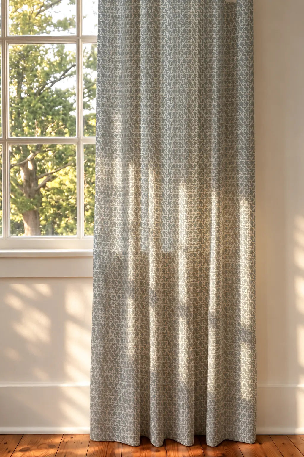 A floor-length curtain panel with a small repeating geometric pattern in muted blue and beige, hanging in front of a window with sunlight and tree shadows. Emily Daws Carolina Rice Fabric in Chambray