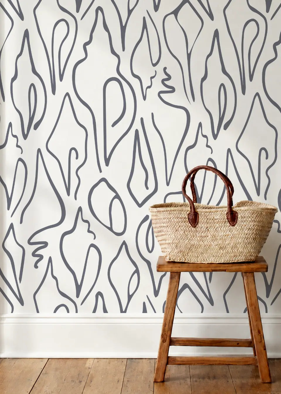 A styled interior scene showing a woven straw tote bag with brown leather handles resting on a small wooden stool against a wall with abstract shell art wallpaper by Emily Daws and a wood floor.  Edisto wallpaper in High Tide