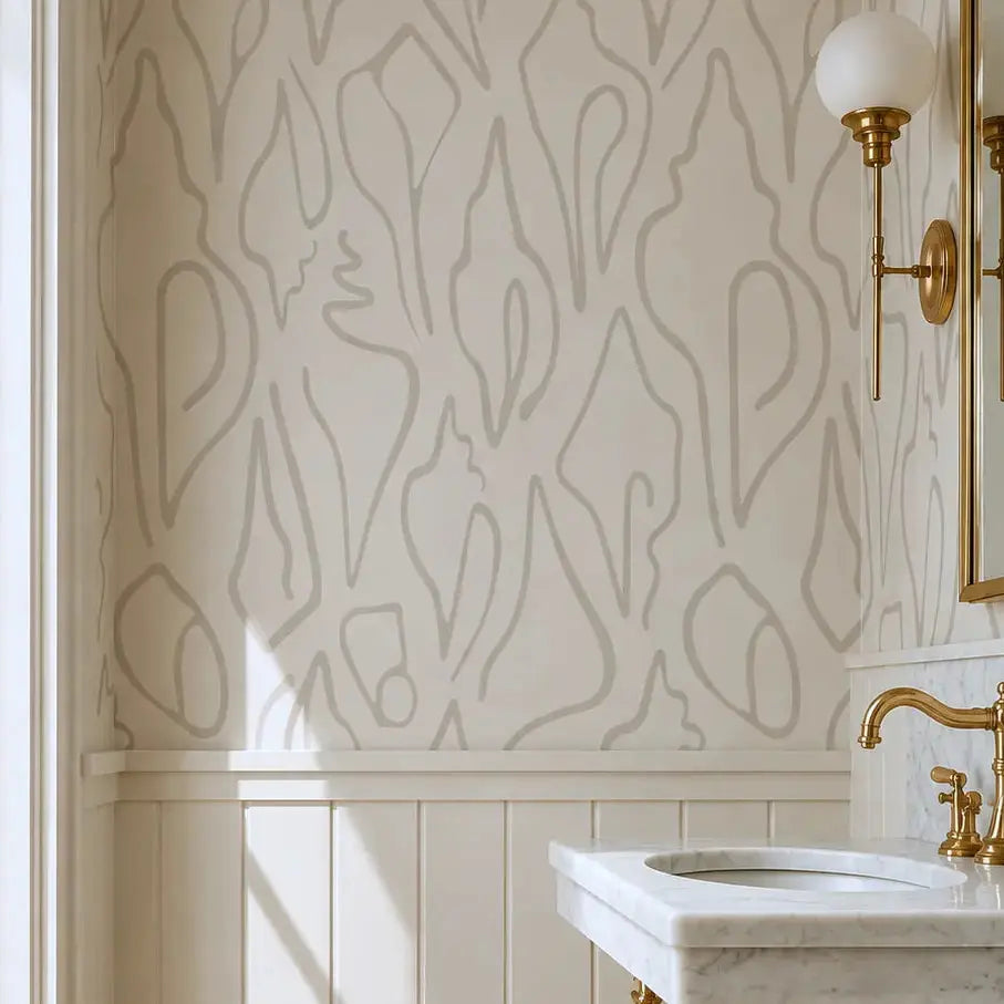 A styled bathroom corner featuring a white marble sink with a polished brass stand, a wall-mounted brass sconce, and a wall with elegant Edisto Wallpaper in beige and white, aabstract shell wallpaper by Emily Daws. 