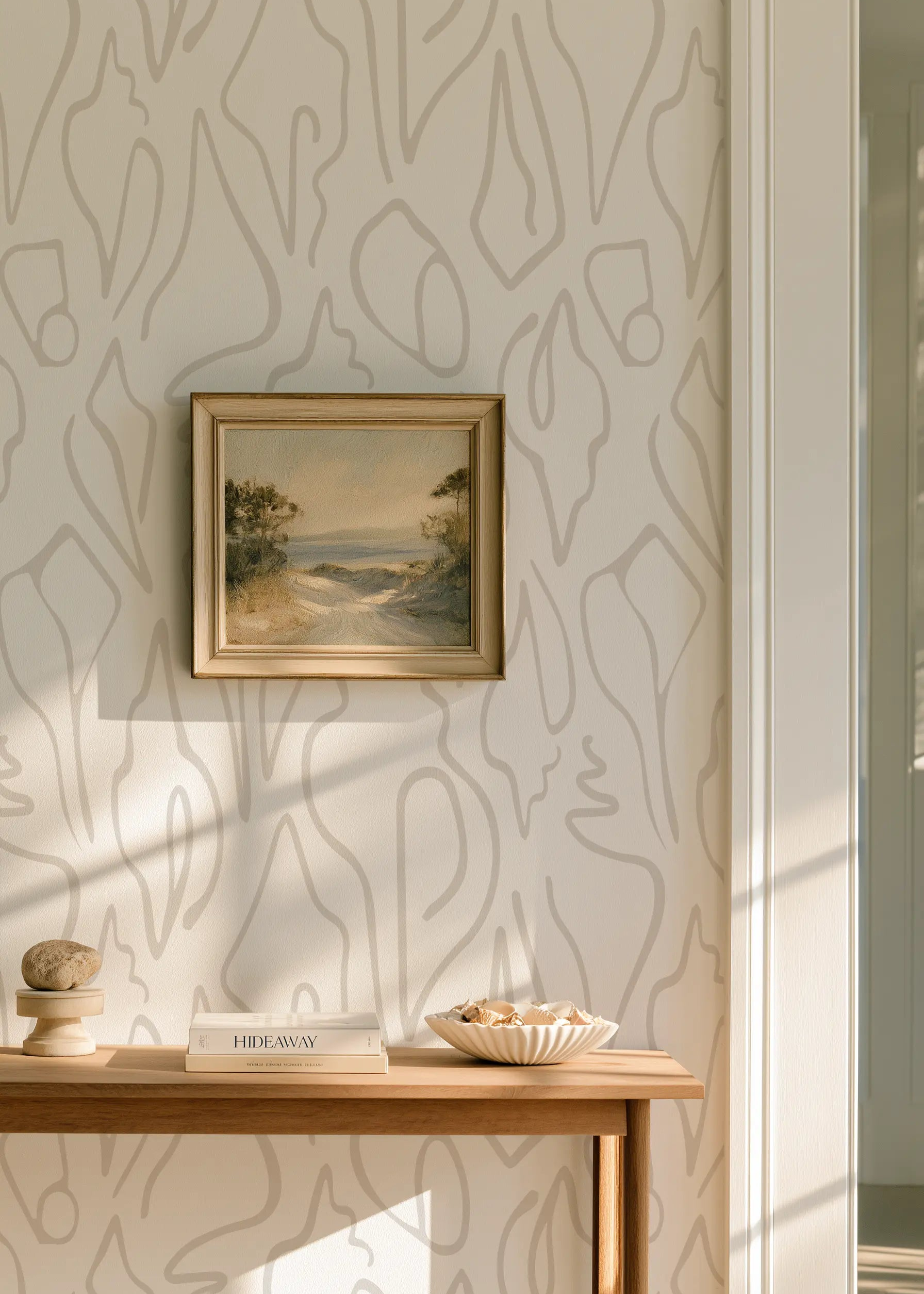 A styled interior corner featuring a small framed landscape painting hanging on a wall with abstract beige shell wallpaper by Emily Daws in Charleston. Below the painting is a light wood console table holding a decorative bowl, stacked books, and a small stone object.  Edisto wallpaper in Shore.