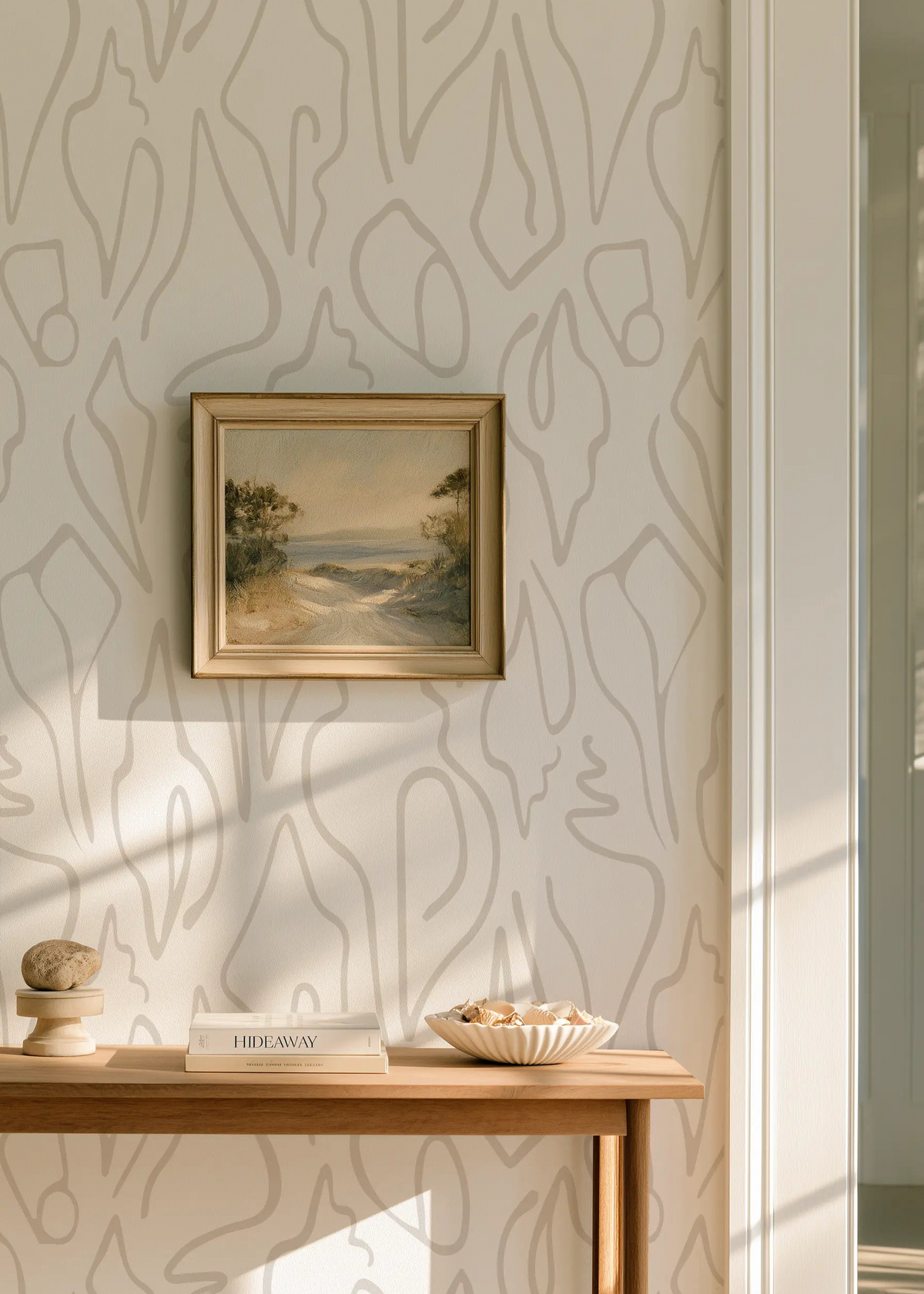 A styled interior corner featuring a small framed landscape painting hanging on a wall with abstract beige shell wallpaper by Emily Daws in Charleston. Below the painting is a light wood console table holding a decorative bowl, stacked books, and a small stone object.  Edisto wallpaper in Shore.