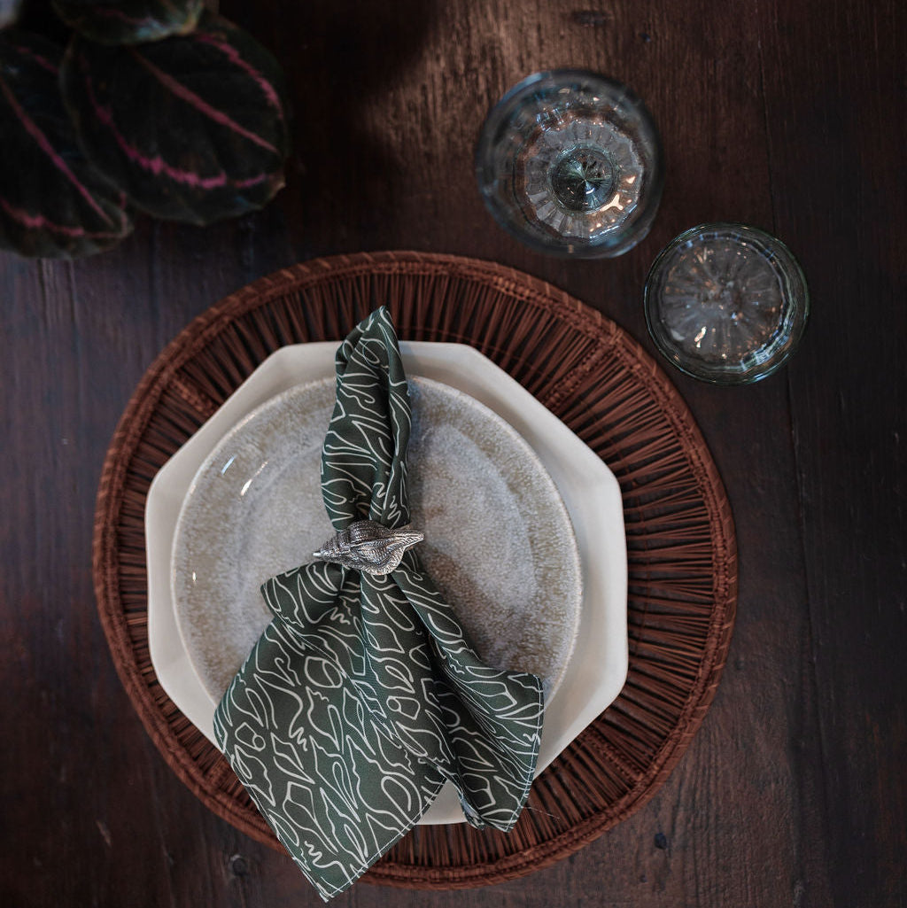 Table setting with a woven charger, white plate, and green napkin on a dark wooden surface.