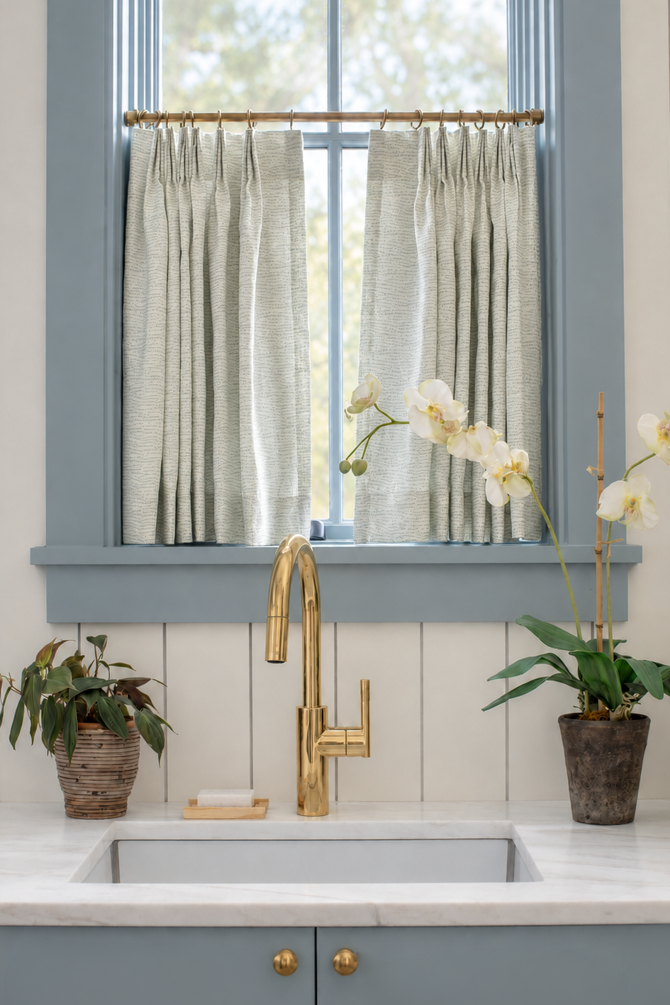 A styled kitchen scene featuring light sage green fabric by Emily Daws in pleated curtain panels hanging on a brass rod over a window with blue trim, above a white shiplap wall, white marble countertop, and a gold faucet.  Harbor Fabric in Celadon by Emily Daws for Southern Living Magazine.