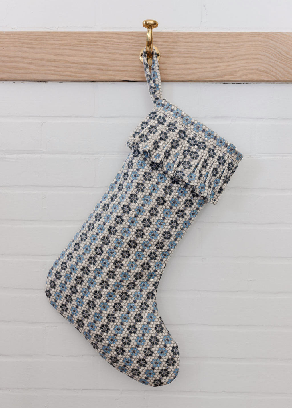 A woven holiday stocking in the Nectar pattern by Emily Daws displayed on a brass hook against white shiplap.