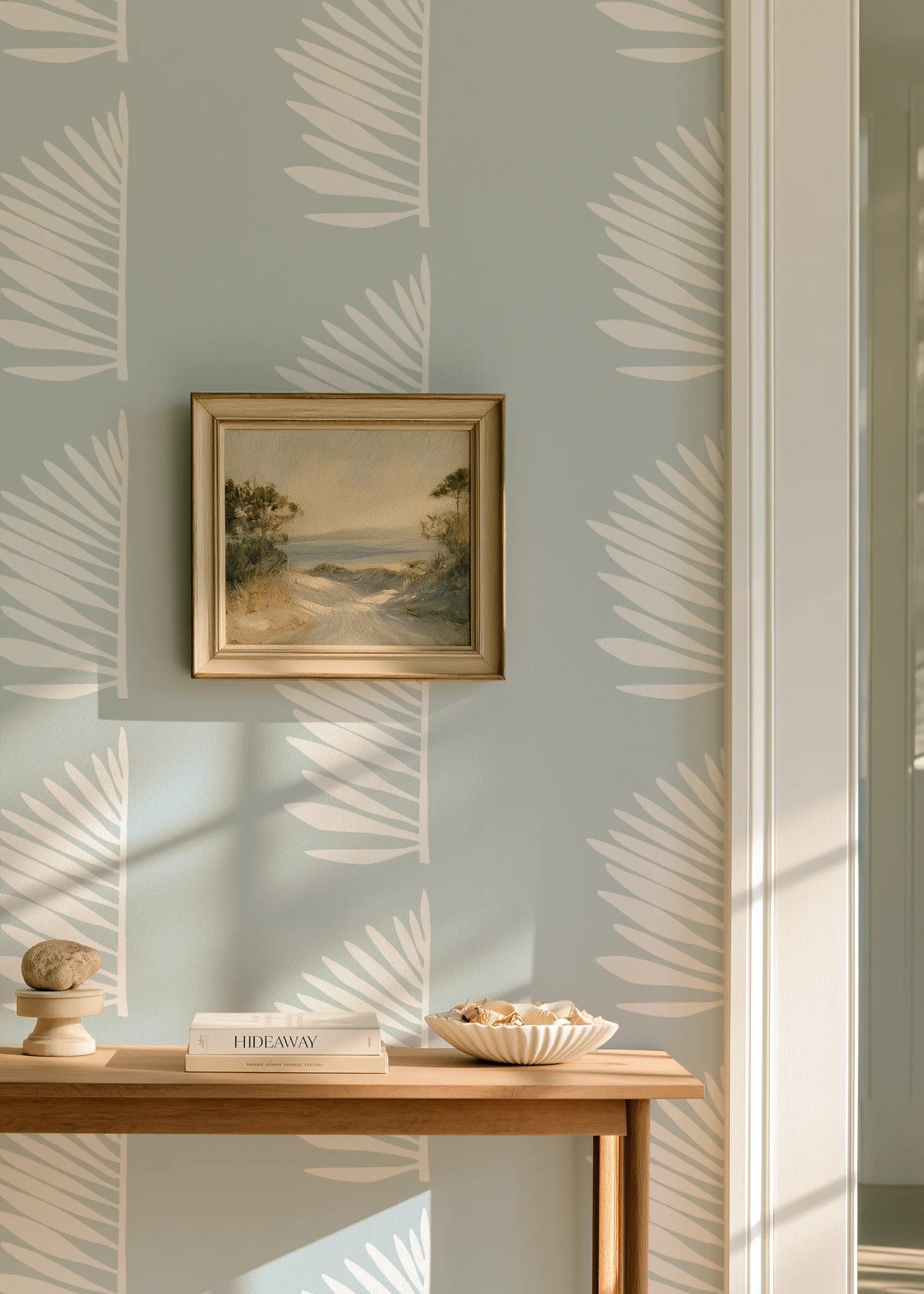 A styled interior scene with pale blue wallpaper featuring a repeating white palm leaf pattern, Palmetto by Emily Daws, a small framed landscape painting on the wall, a wooden console table, books, and a bowl of fruit in warm natural light.  Palmetto wallpaper  in Haint Blue, designed in Charleston.
