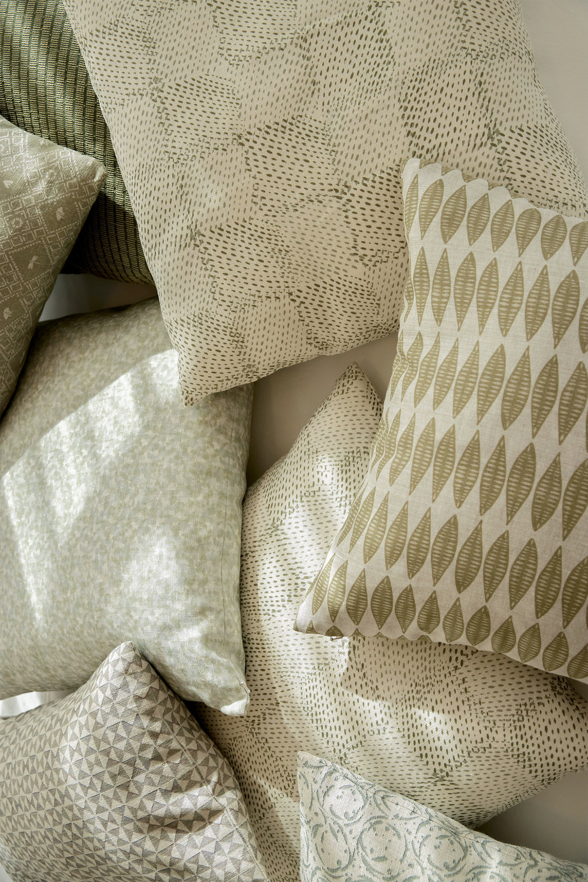 A collection of decorative throw pillows in neutral tones, featuring various geometric, textured, and leaf-like patterns in beige, cream, and muted olive green. Emily Daws Belgian Linen throw pillows.
