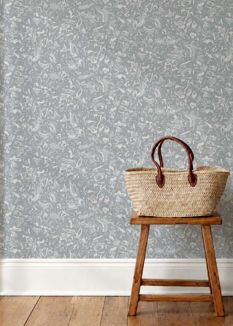 A woven straw tote bag with dark brown leather handles sits on a small wooden stool against a wall covered in light gray and white abstract patterned wallpaper, with a light wood floor below.