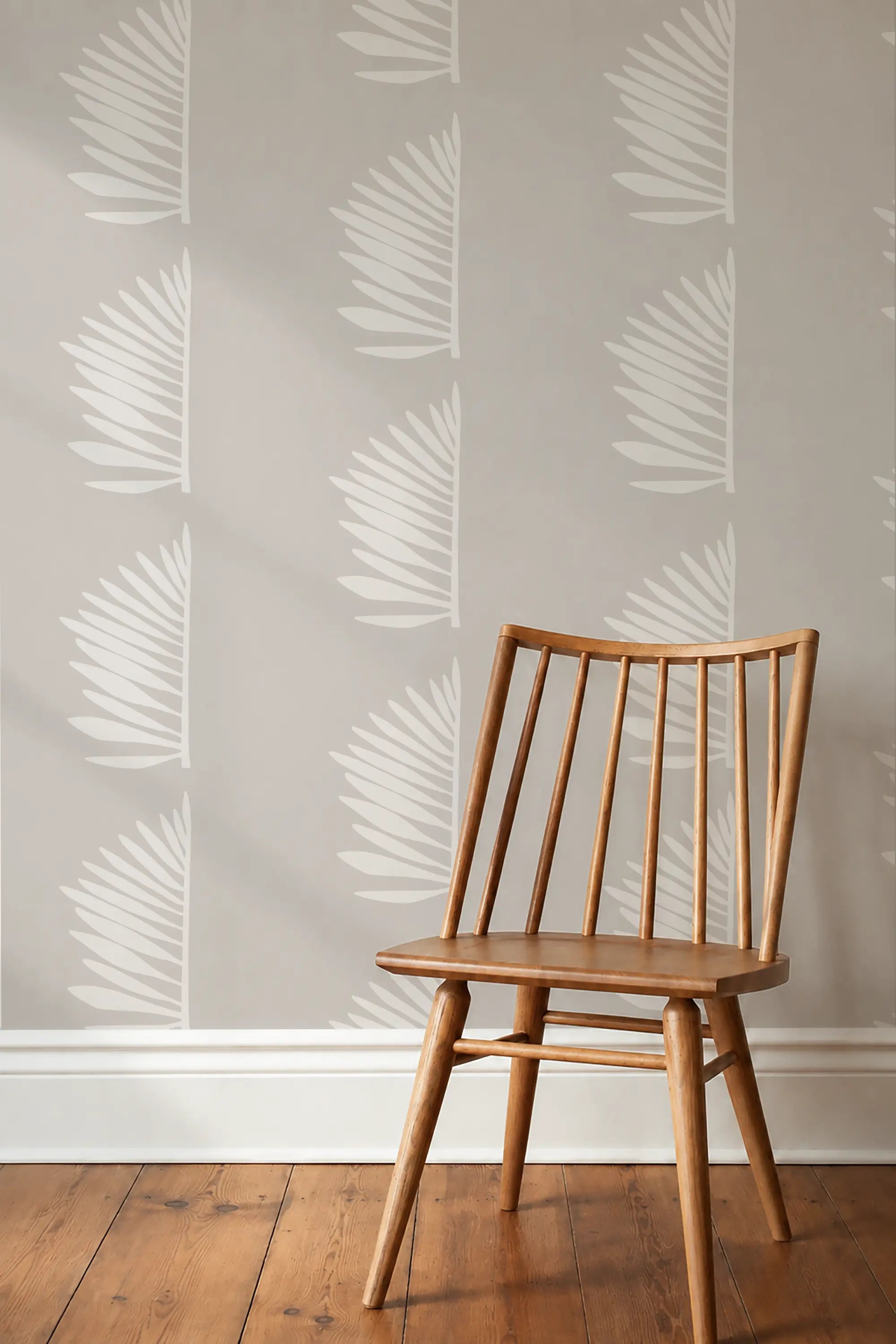 A light natural wood spindle-back chair positioned in front of a wall with repeating white palm leaf decals on a warm gray background, set on a medium brown wooden floor. Palmetto wallpaper by Emily Daws