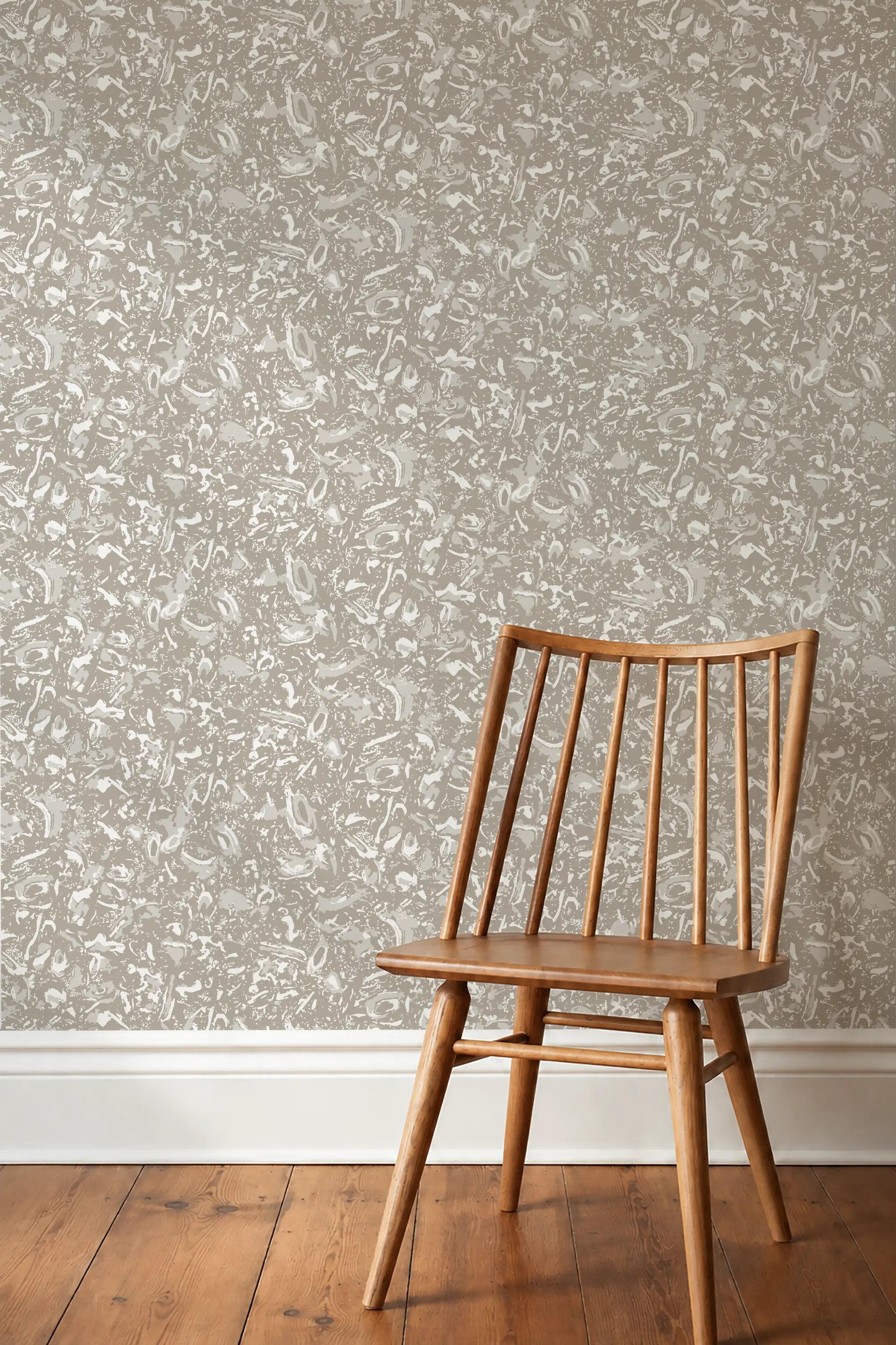 A wooden spindle-back chair placed in front of a wall covered in a light beige and white shell patterned wallpaper by Emily Daw, with a medium-brown wood floor visible below.  River Reef Wallpaper in Natural