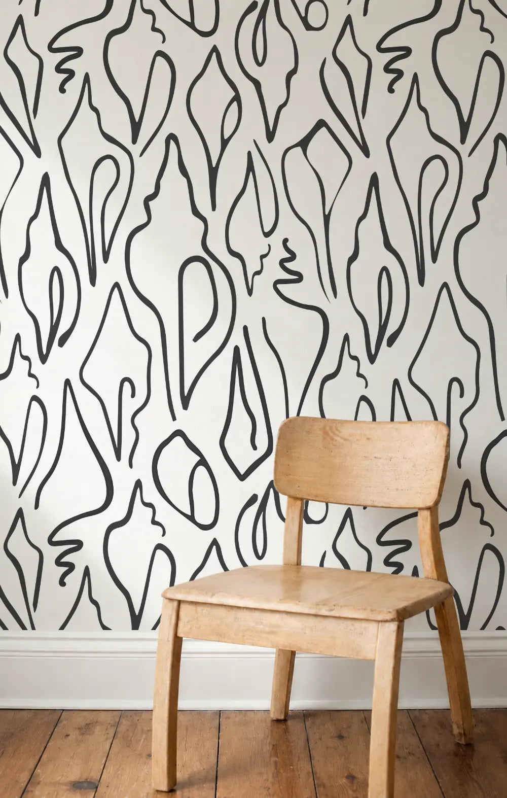 A light natural wood chair placed in front of a white wall covered in black abstract shell-patterned wallpaper by Emily Daws, with a wooden floor and white baseboard visible.  Edisto wallpaper in Jet.