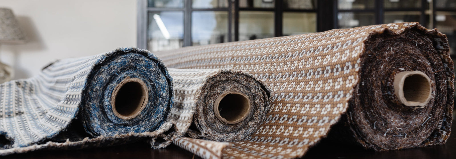 Three rolls of Nectar woven fabric on a wooden surface with a blurred background of Emily Daws Showroom.