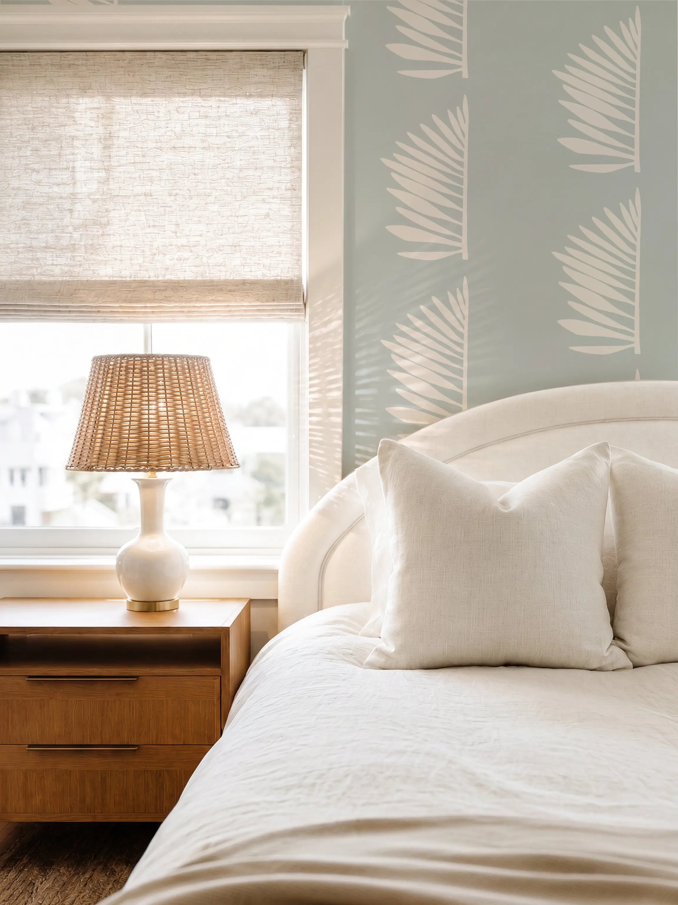 A styled bedroom corner featuring a white bed with textured cream bedding, a wooden nightstand with a woven lamp, and a light blue wall with white Palmetto leaf wallpaper by Emily Daws