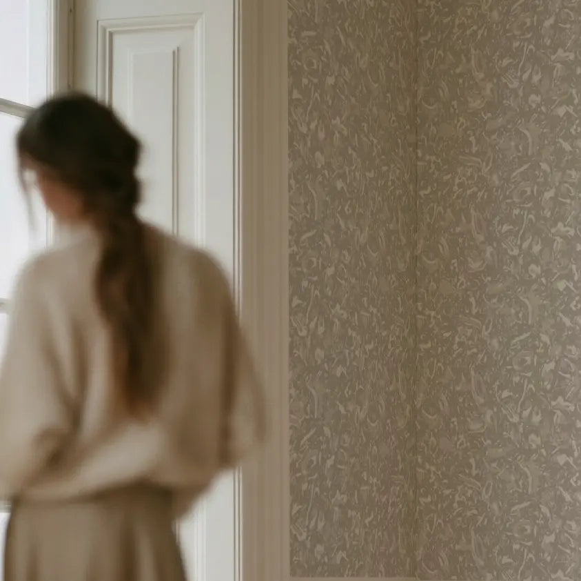 A woman in a cream-colored sweater and skirt stands in profile near a window, looking at a wall covered in textured beige and gray shell-patterned wallpaper by Emily Daws. River Reef wallpaper detail in Pearl