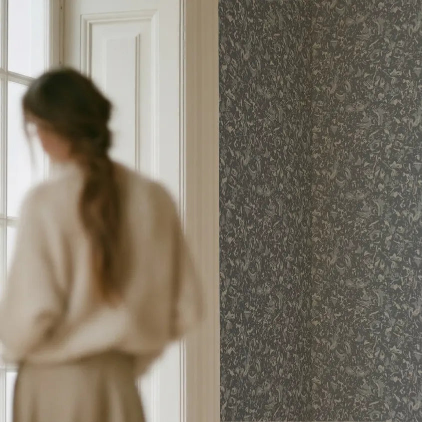 A woman in a cream sweater stands near a window, looking at a wall covered in blue and beige shell-patterned wallpaper by Emily Daws