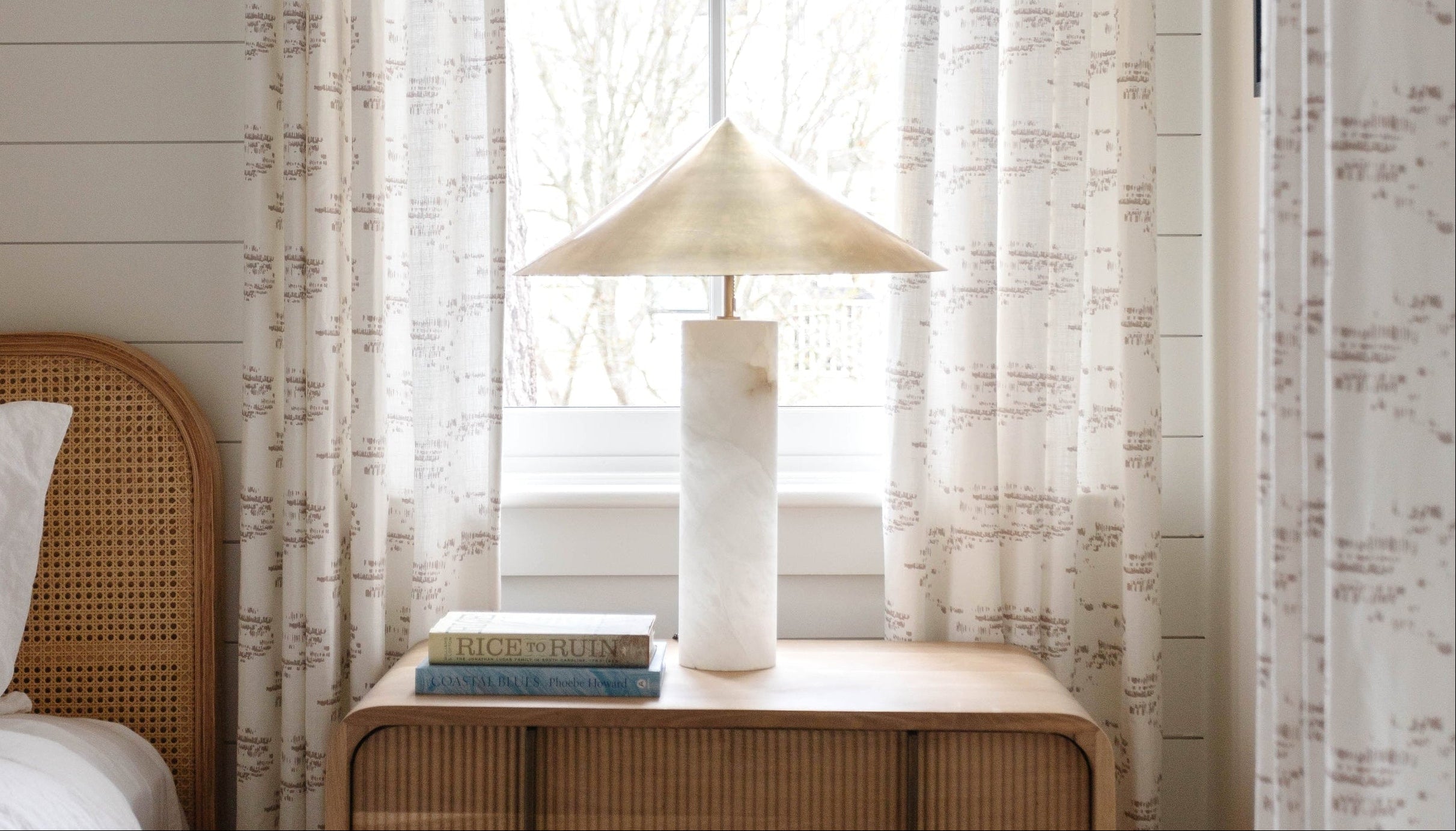 Nightstand with lamp and books next to a bed in a bedroom with patterned curtains by Emily Daws.