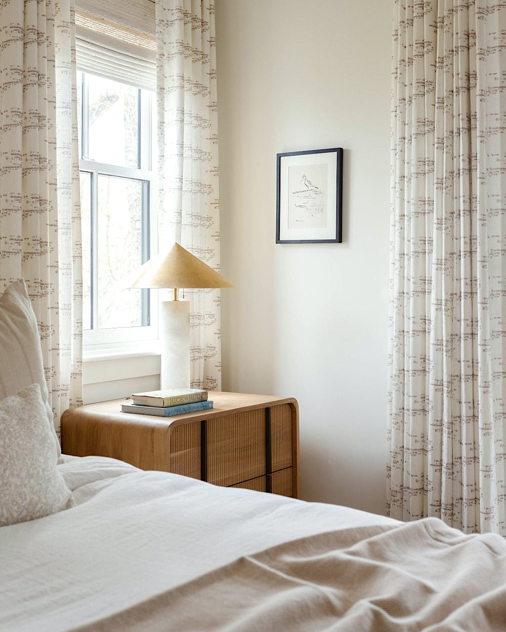 Nightstand with lamp and books beside a bed with patterned curtains by Emily Daws.